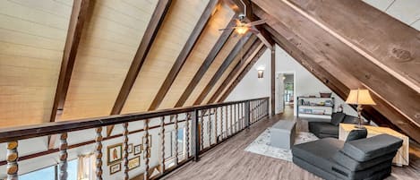 An attic room with exposed wooden beams, a wooden railing, a sofa, a lamp, and a view of the living area.