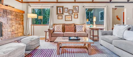 A cozy living room with a stone fireplace, a sofa set, a coffee table, and a view of the outdoors through large windows.