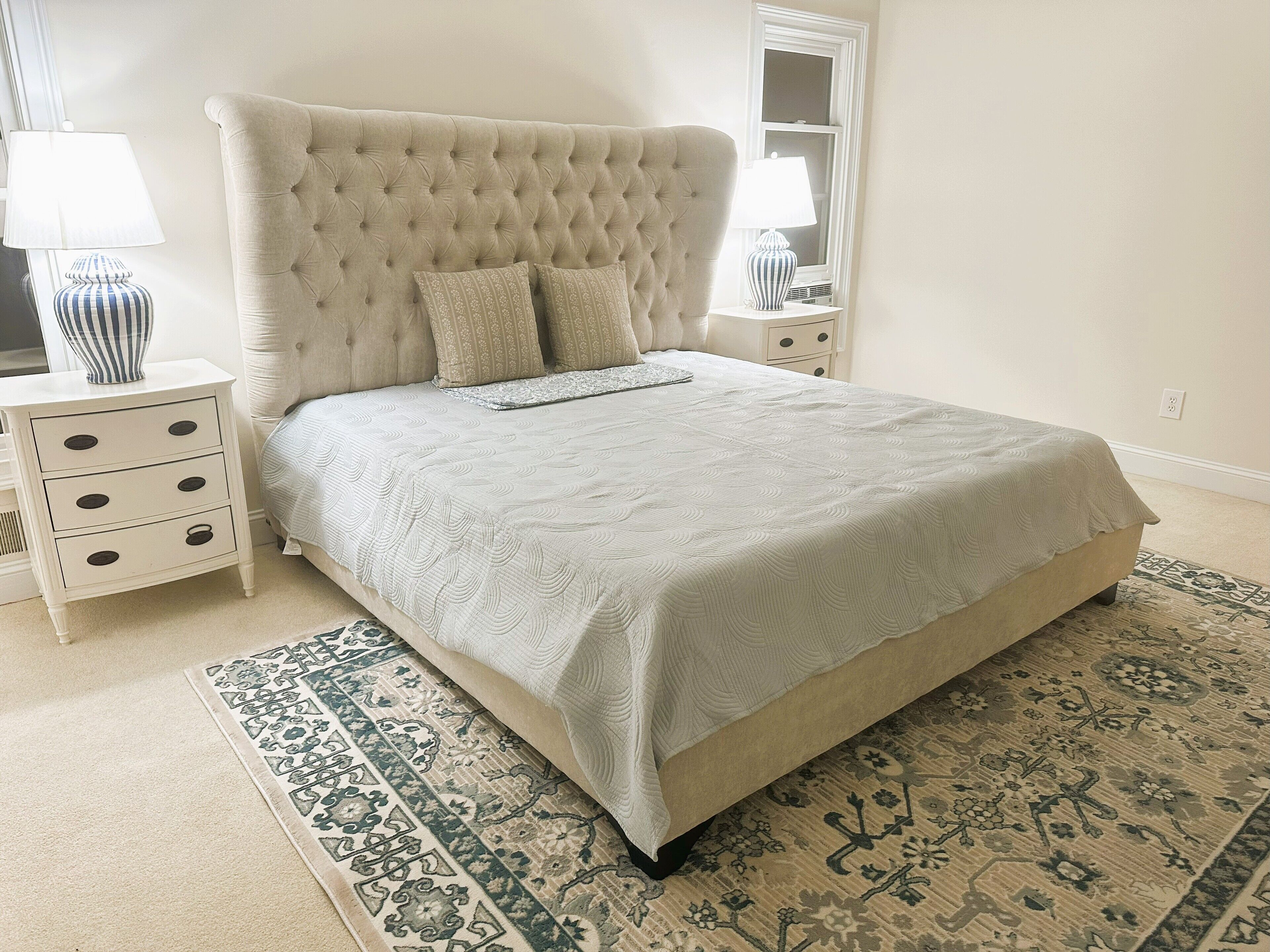 A bedroom with a tufted headboard, a large bed with a patterned bedspread, two bedside tables with lamps, and a decorative rug.