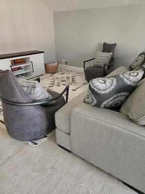 A living room with a grey sofa, a grey armchair, and a patterned rug. There is a white cabinet with books and a basket. - Charming 4-bedroom house, 4-king beds, Extended + Corporate Stays, Near Airport (Fayetteville)