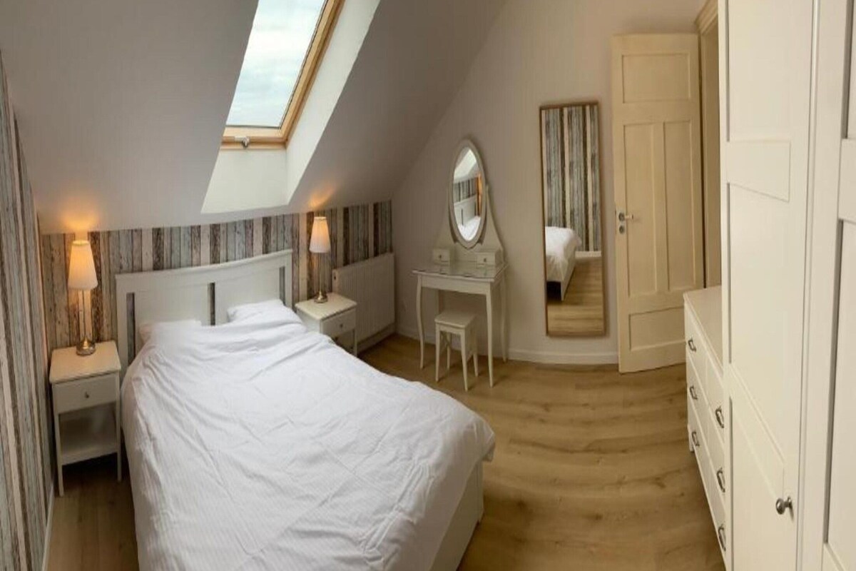 Ein Schlafzimmer mit einem großen Bett, einem Schreibtisch mit Spiegel, einem Kleiderschrank und einem Dachfenster.