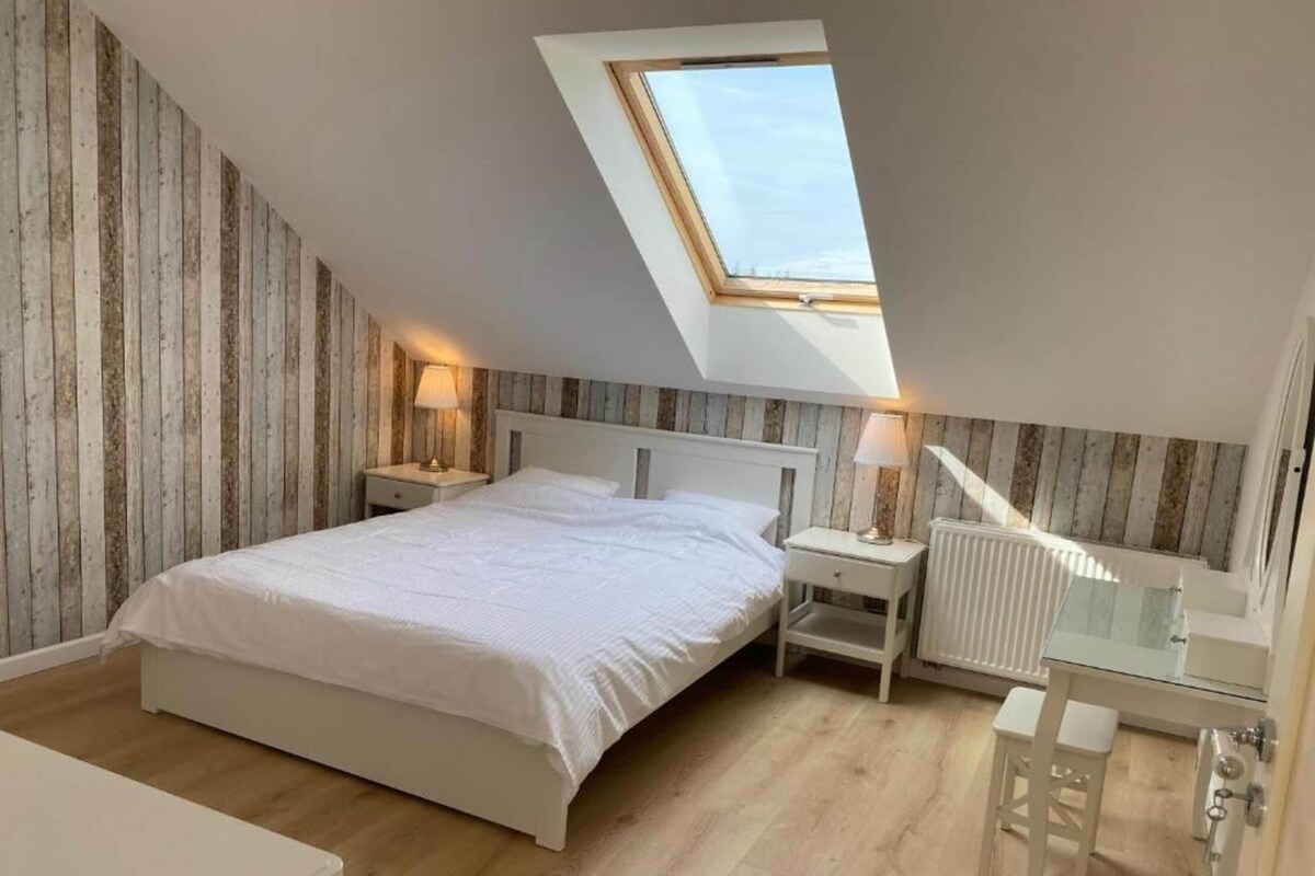 Ein Schlafzimmer mit schrägem Dach, einem Oberlicht, einem Bett, Nachttischen, einem Heizkörper und einem kleinen Hocker.