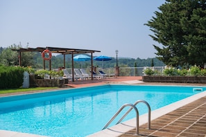 A swimming pool with lounge chairs and umbrellas, surrounded by greenery and a view of hills. - Apartment in Farmhouse With Garden and Pool (Montaione)