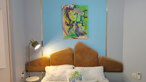 Un dormitorio con cama, cabecero, lámpara de noche, un cuadro en la pared, un cubo de basura verde y un enchufe.