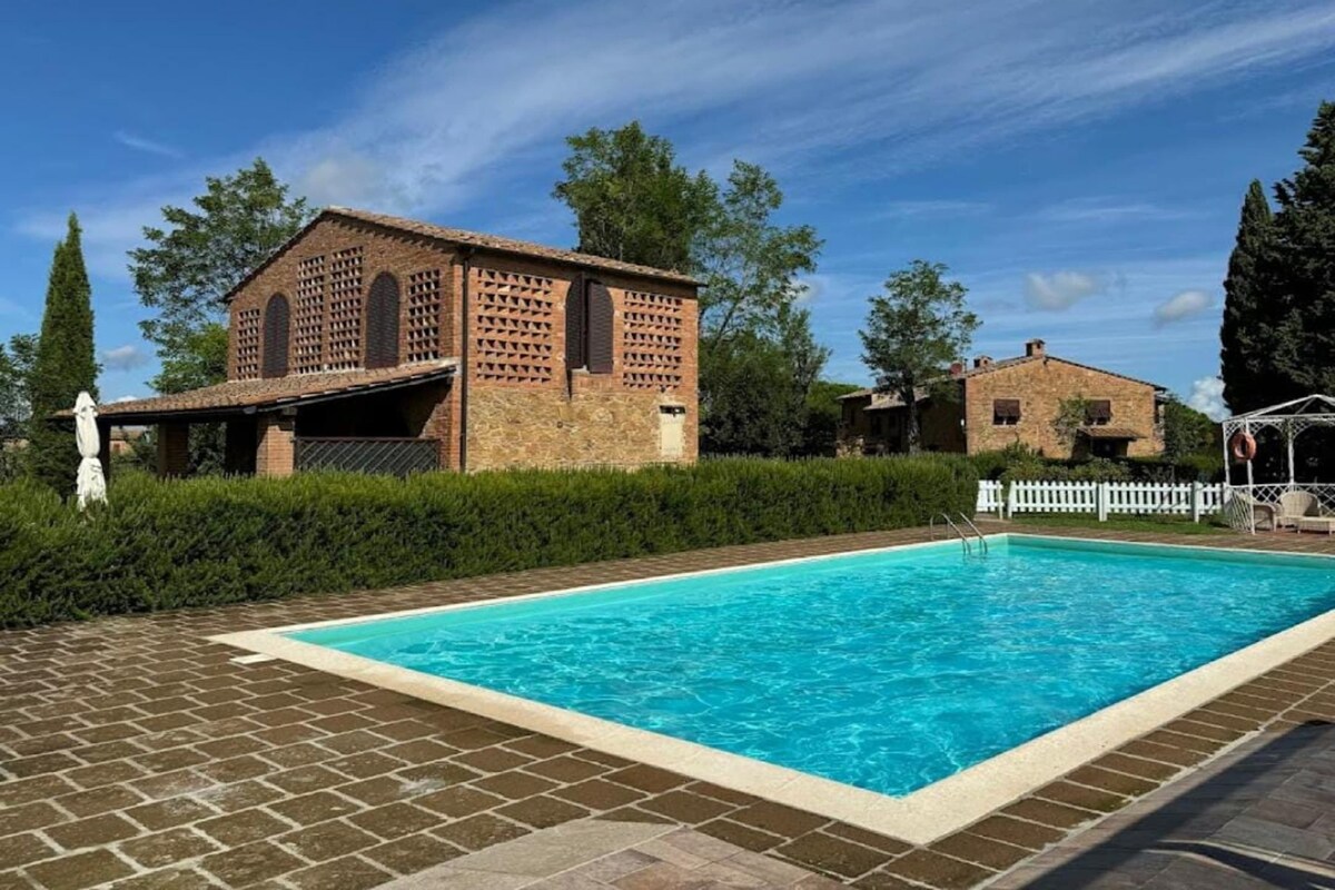 A swimming pool with a brick border, a brick building with arched windows, and a white fence.