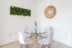 A dining area with a glass table, white chairs, a decorative wall hanging, and a mirror. - Apartment 'Mistral Apartment La Manga' with Sea View, Wi-Fi and Air Conditioning (San Javier)