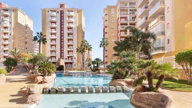 A swimming pool with a dolphin design, surrounded by palm trees and large rocks, with high-rise buildings in the background. - Apartment 'Mistral Apartment La Manga' with Sea View, Wi-Fi and Air Conditioning (San Javier)