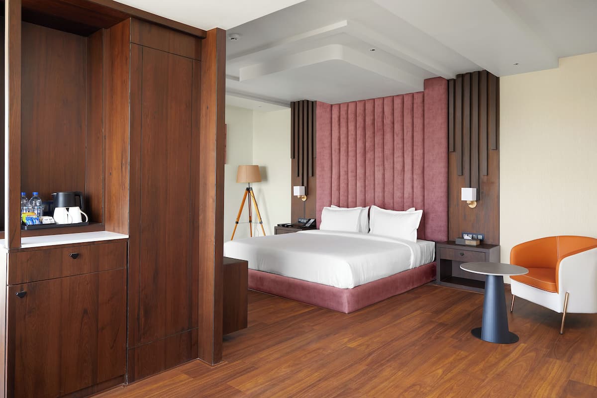 A modern hotel room with a wooden wardrobe, a large bed with white bedding, a bedside table with a lamp, and a small orange and white chair.