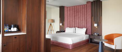 A modern hotel room with a wooden wardrobe, a large bed with white bedding, a bedside table with a lamp, and a small orange and white chair.