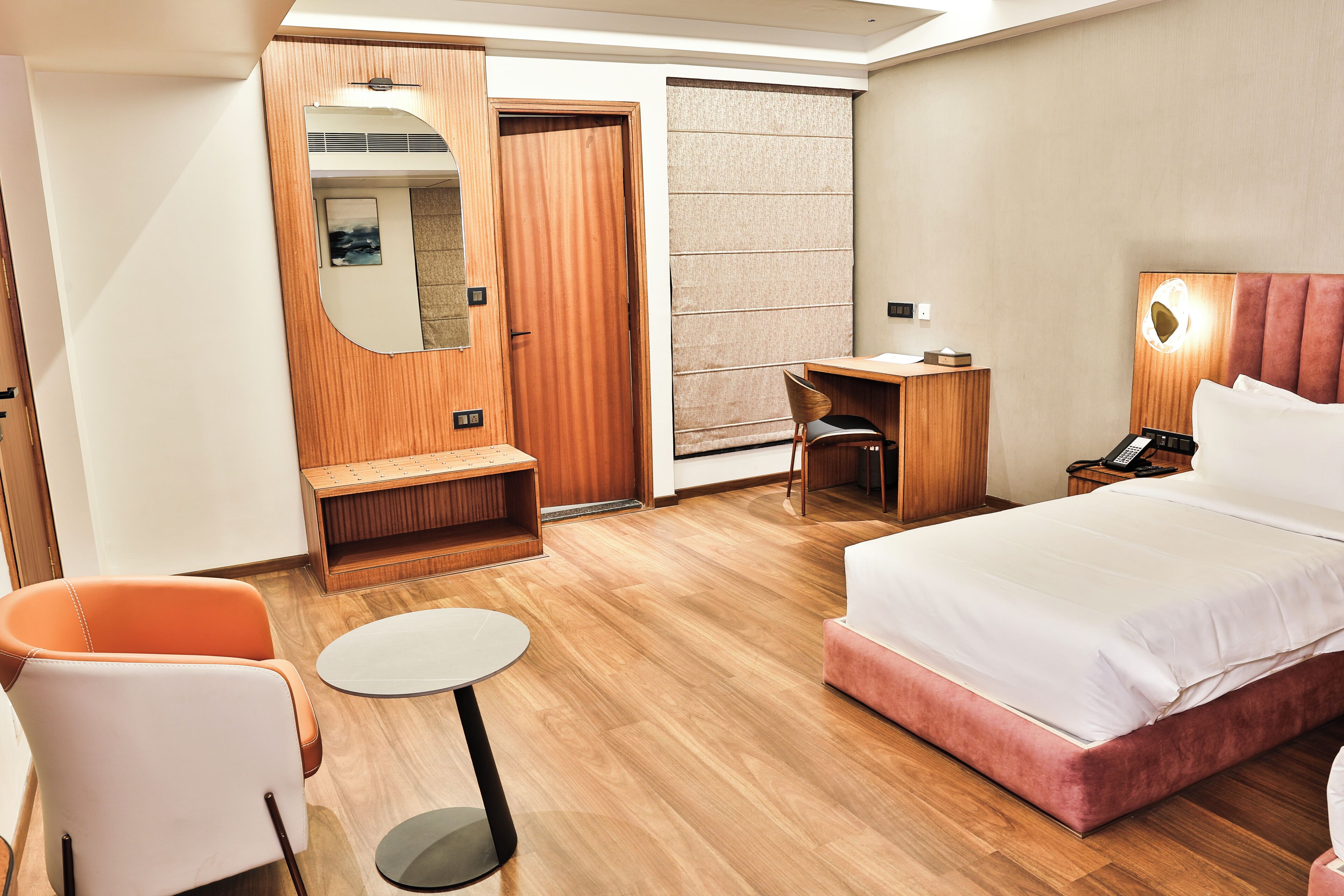 A hotel room with a bed, a desk, a chair, a small table, a mirror, and a wardrobe.