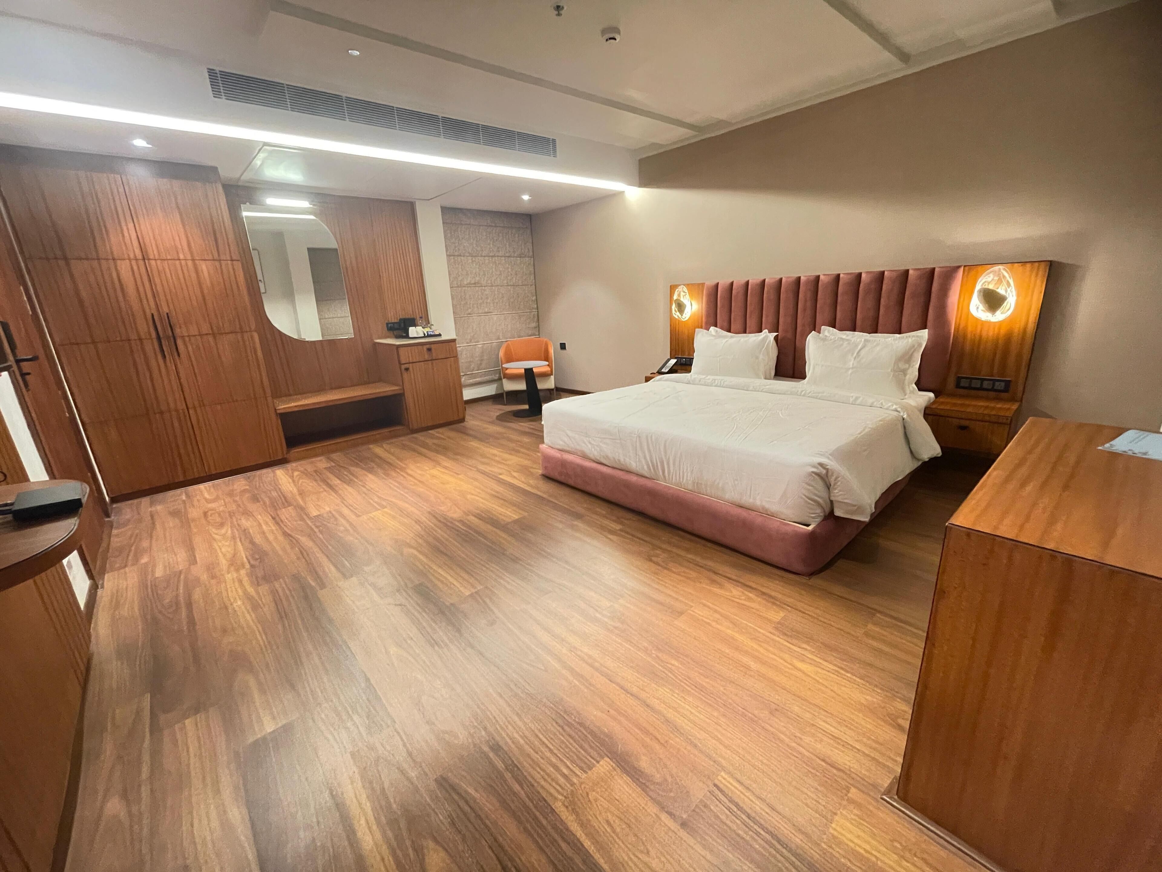 A modern hotel room with a large bed, wooden flooring, a wardrobe, a desk, and a chair.