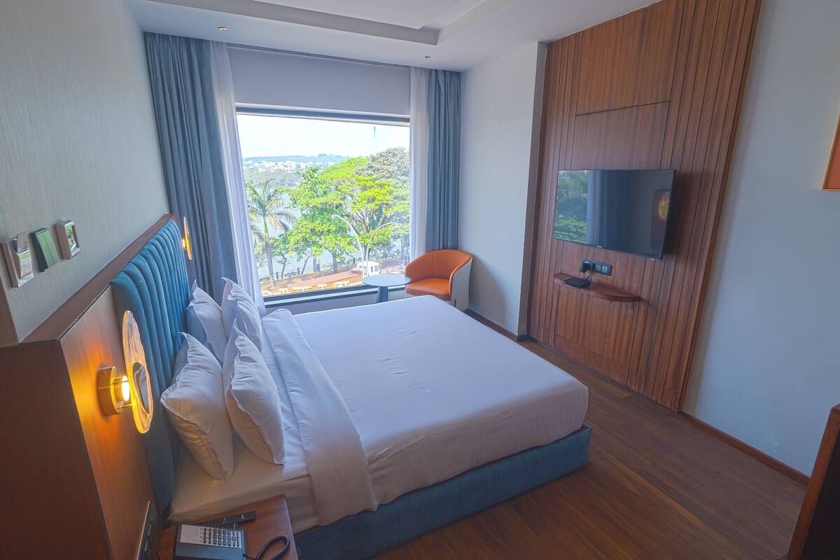 A hotel room with a large bed, a TV mounted on the wall, a desk with a phone, and a view of the outdoors through a window.
