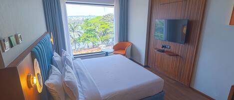 A hotel room with a large bed, a TV mounted on the wall, a desk with a phone, and a view of the outdoors through a window.