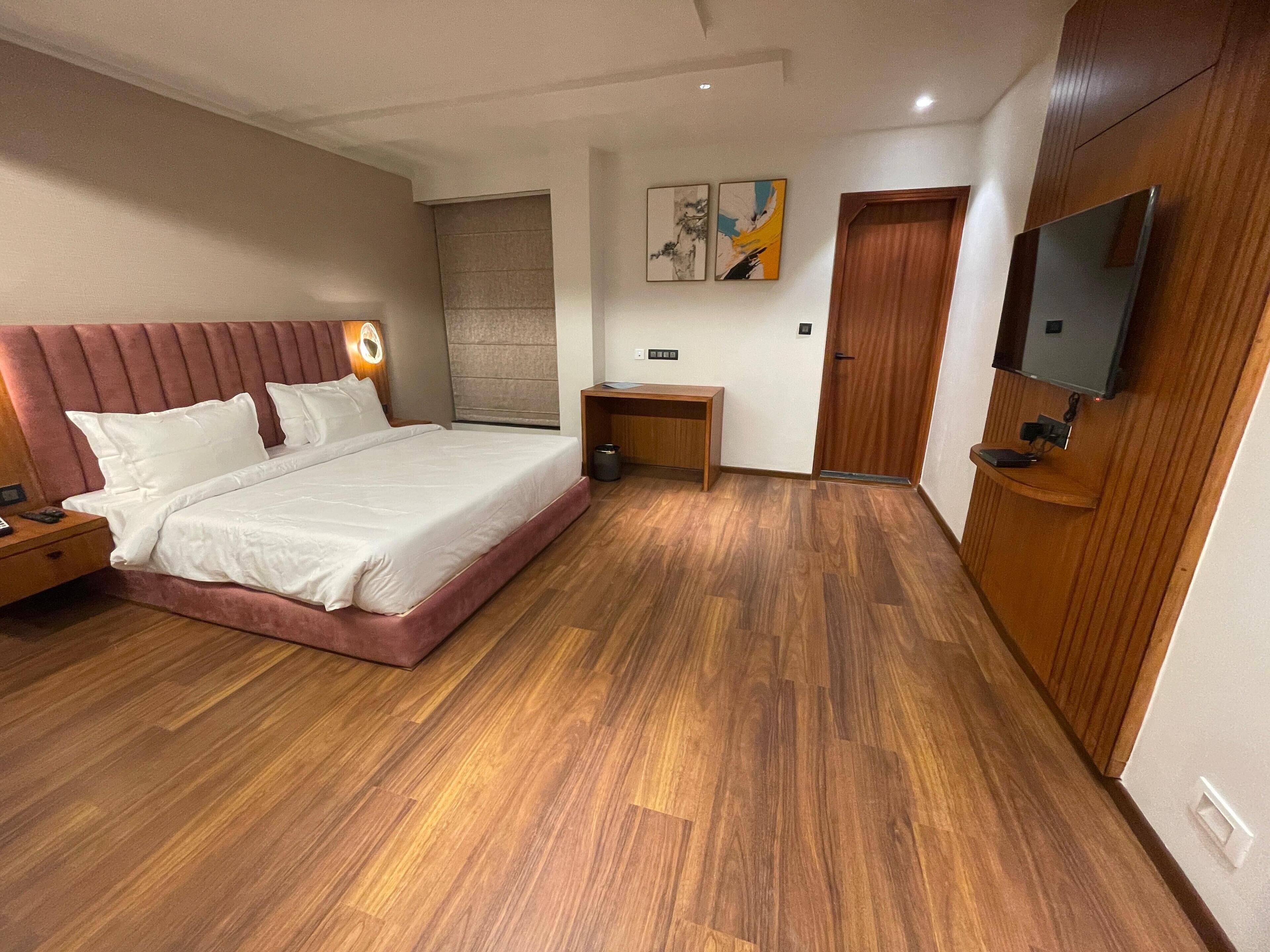 Moderni hotellihuone, jossa on suuri sänky, puulattia, puinen sängynpääty, yöpöytä, pieni sivupöytä, työpöytä, tuoli, seinälle asennettu televisio ja ovi.
