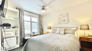 A bedroom with a bed, bedside lamps, a ceiling fan, a window with curtains, a wall-mounted artwork, and a shelf with a microwave and other items. - Canmore Windtower Hotel Room  (Canmore)