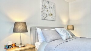 A neatly made bed with a grey headboard, two bedside lamps, a small wooden nightstand with a tissue box, and a wall-mounted abstract floral artwork. - Canmore Windtower Hotel Room  (Canmore)