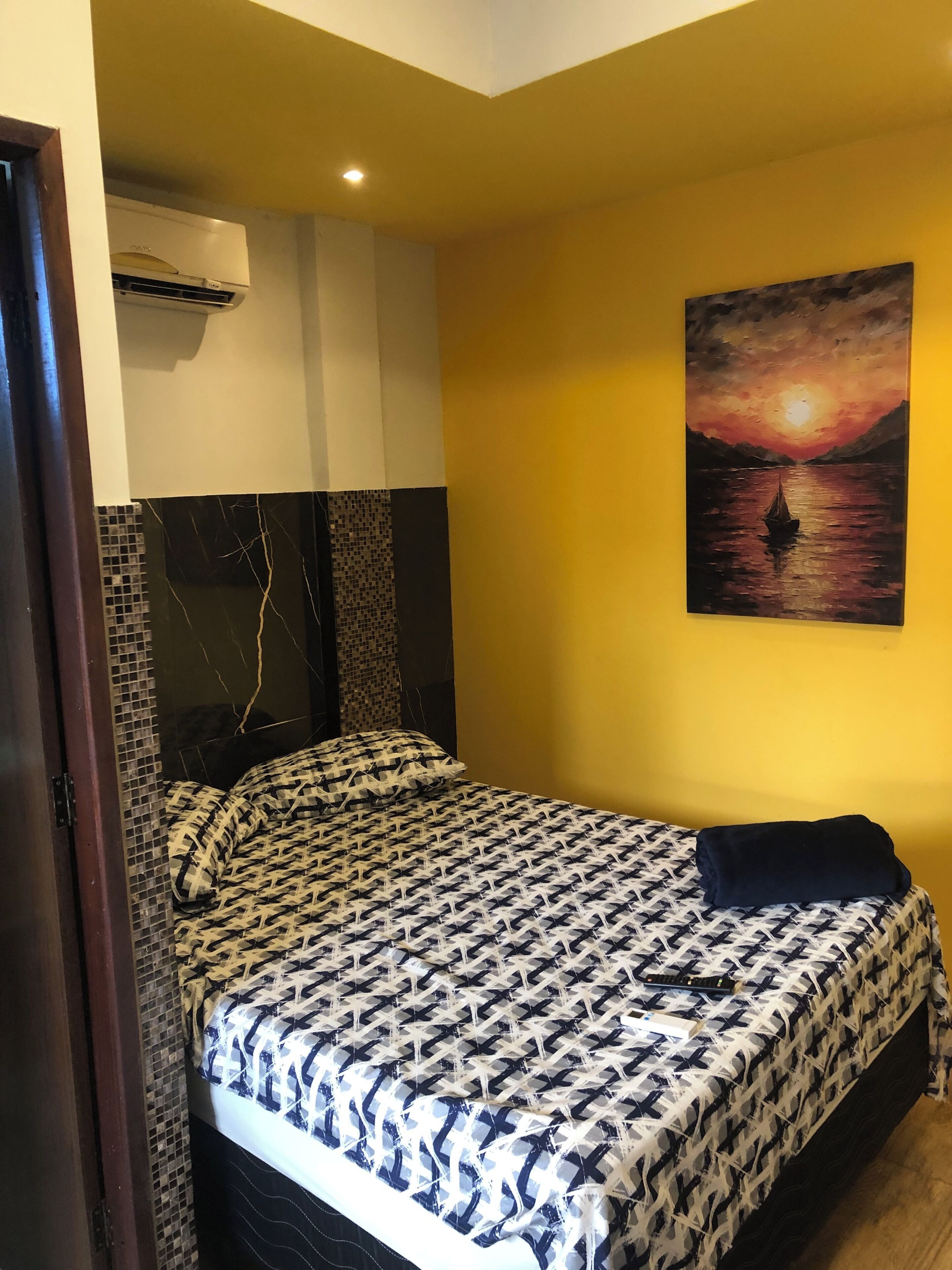 A bedroom with a patterned bedspread, a painting of a sunset over water, and an air conditioning unit above the bed.