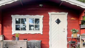 A red wooden cabin with a white-framed window and door, a small bench with a gray blanket, and a potted plant on the porch.