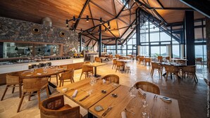 Uma sala de jantar espaçosa com mesas e cadeiras de madeira, uma parede de pedra e grandes janelas que permitem a entrada de luz natural.