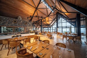 Un comedor amplio con mesas y sillas de madera, pared de piedra y ventanas grandes que dejan entrar luz natural.