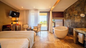 Quarto de hotel com uma cama grande, área de estar com cadeiras, TV, banheira e vista para o exterior.