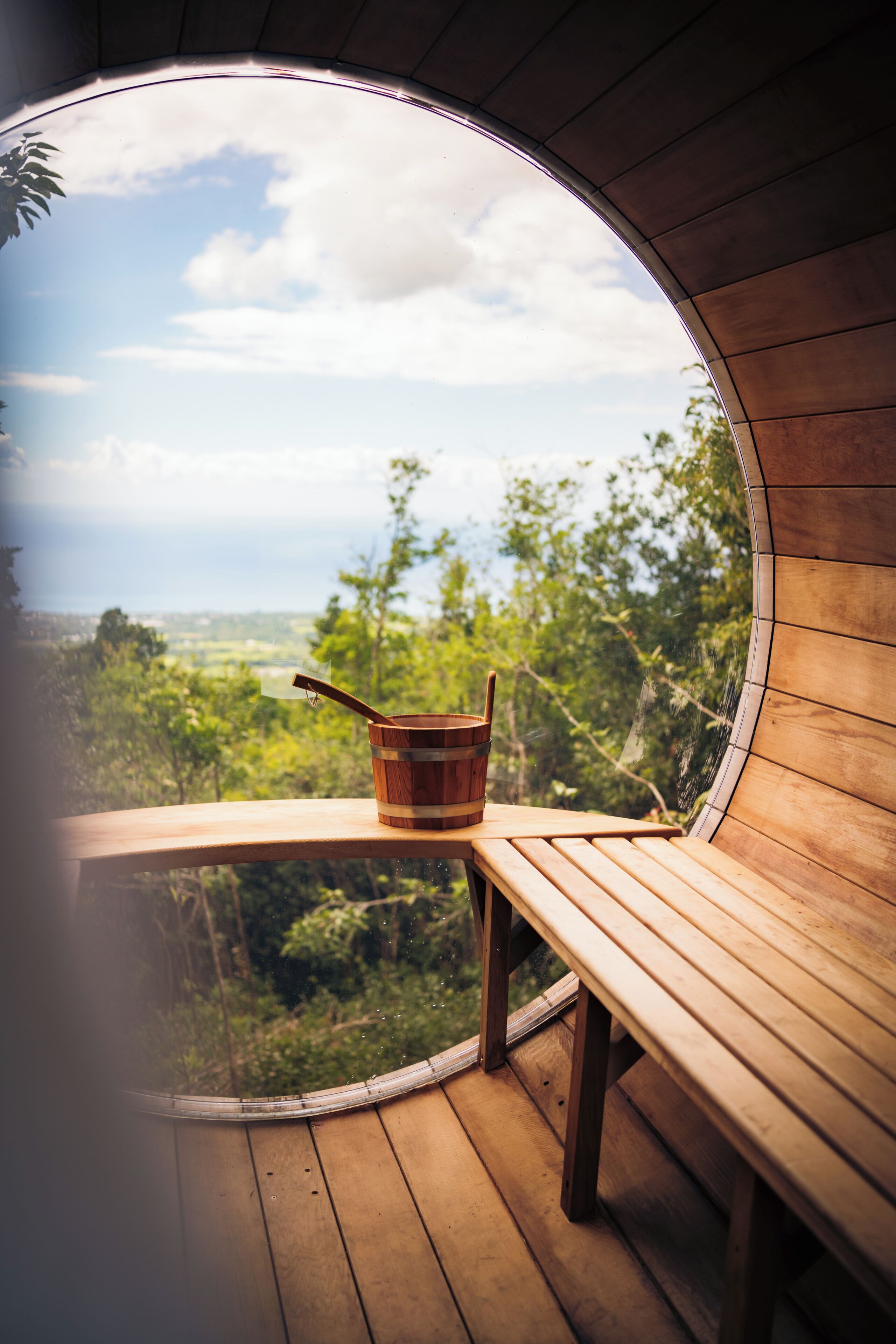 Een houten bankje en een houten emmer op een balkon met een rond raam dat uitkijkt over een groen landschap.