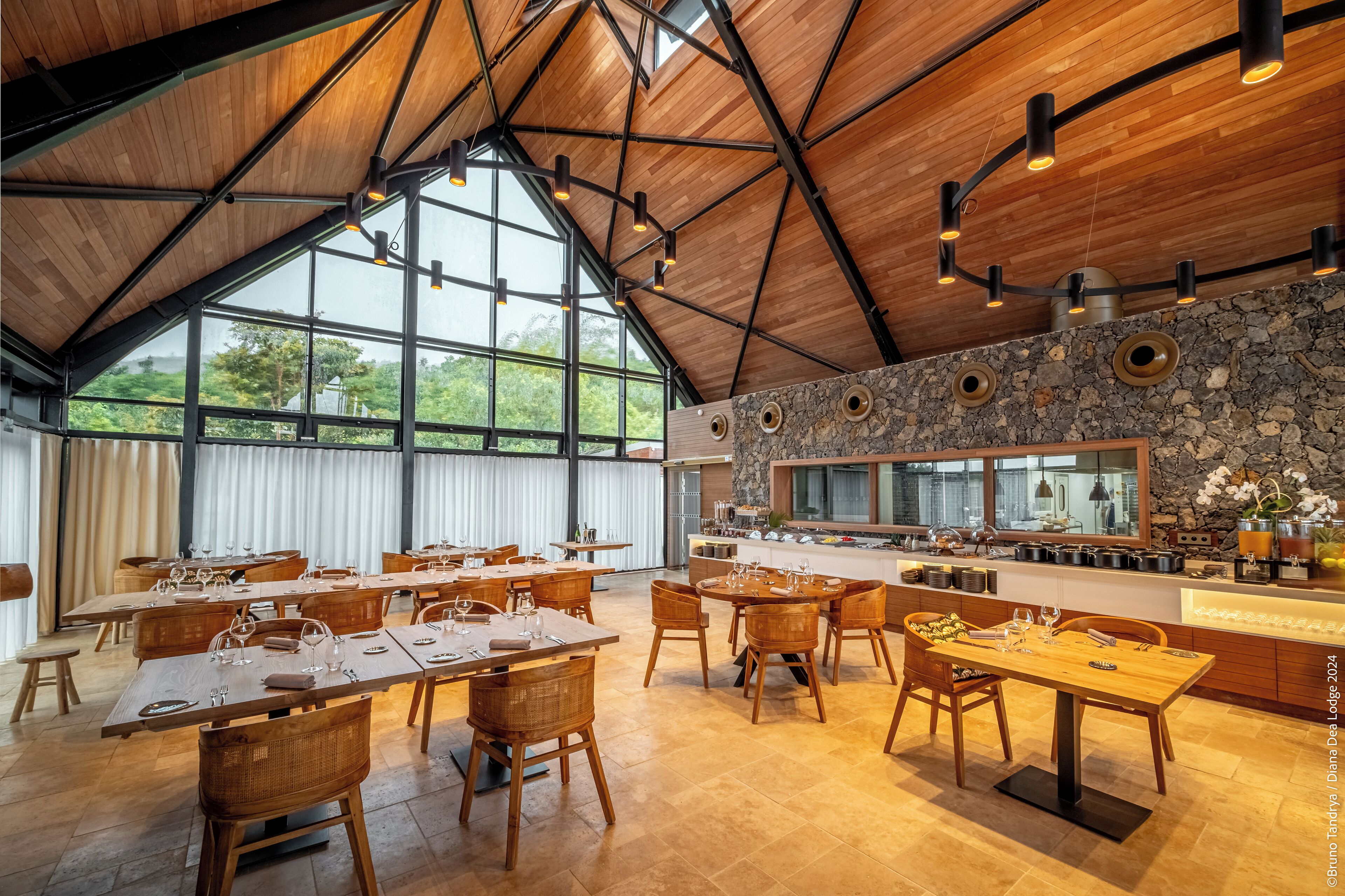 Een ruime eetruimte met hoge plafonds en grote ramen, met houten tafels en stoelen, een accentmuur van steen en een moderne keuken op de achtergrond.