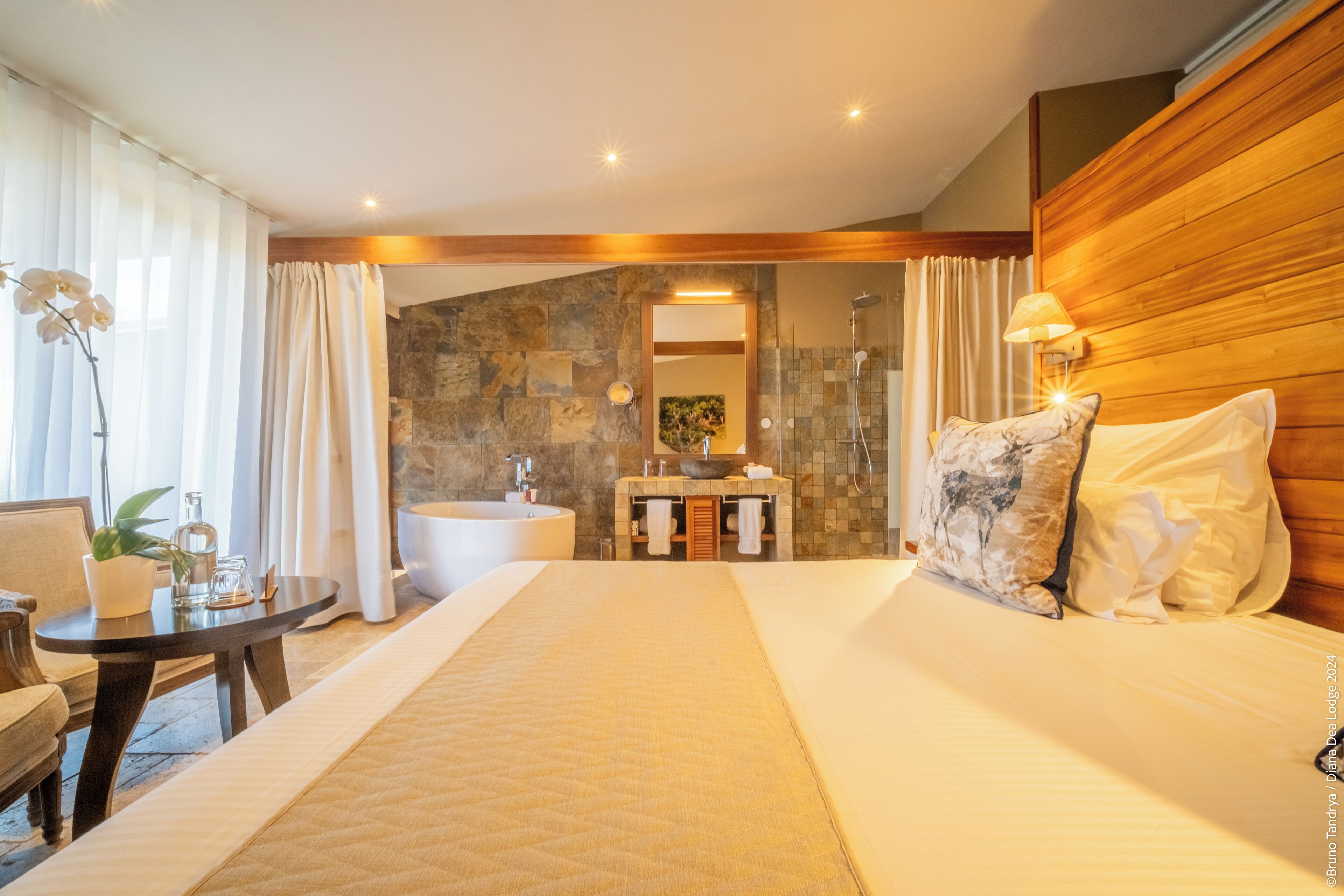 A hotel room with a large bed, a wooden headboard, a sitting area with a table and chairs, a bathroom with a bathtub, and a stone wall.
