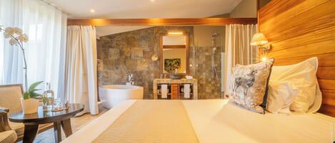 A hotel room with a large bed, a wooden headboard, a sitting area with a table and chairs, a bathroom with a bathtub, and a stone wall.