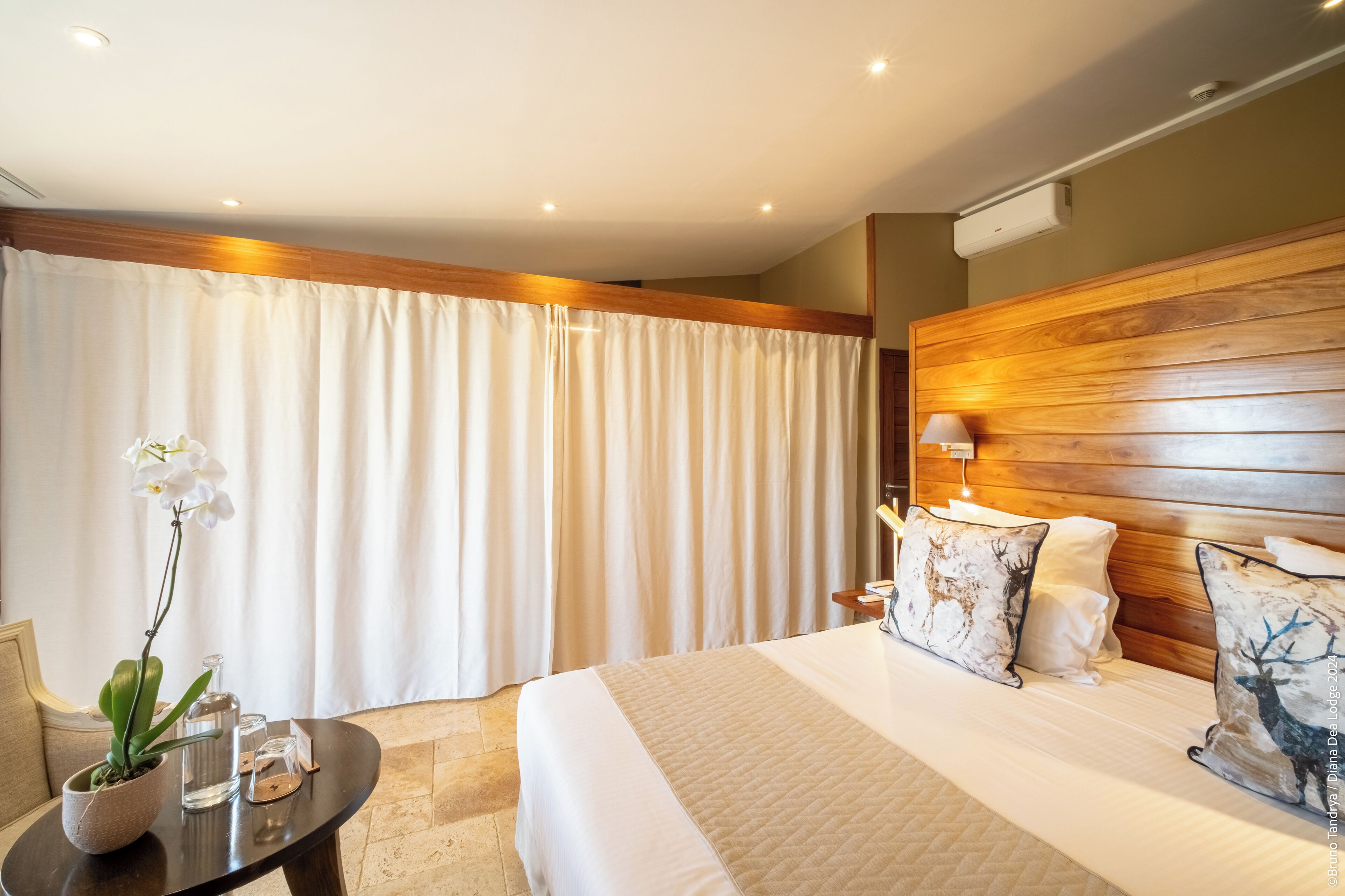 A hotel room with a large bed, wooden headboard, white curtains, a small table with a potted plant, and decorative pillows.
