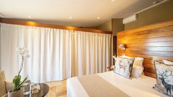 A hotel room with a large bed, wooden headboard, white curtains, a small table with a potted plant, and decorative pillows.