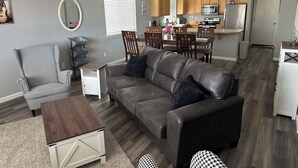 Ein modernes Wohnzimmer mit grauem Sofa, Zwillingskarree-Sessel und Couchtisch aus Holz. Im Hintergrund sind ein Essbereich mit Holzkästen und eine Küche mit Mikrowelle und Kühlschrank zu sehen.