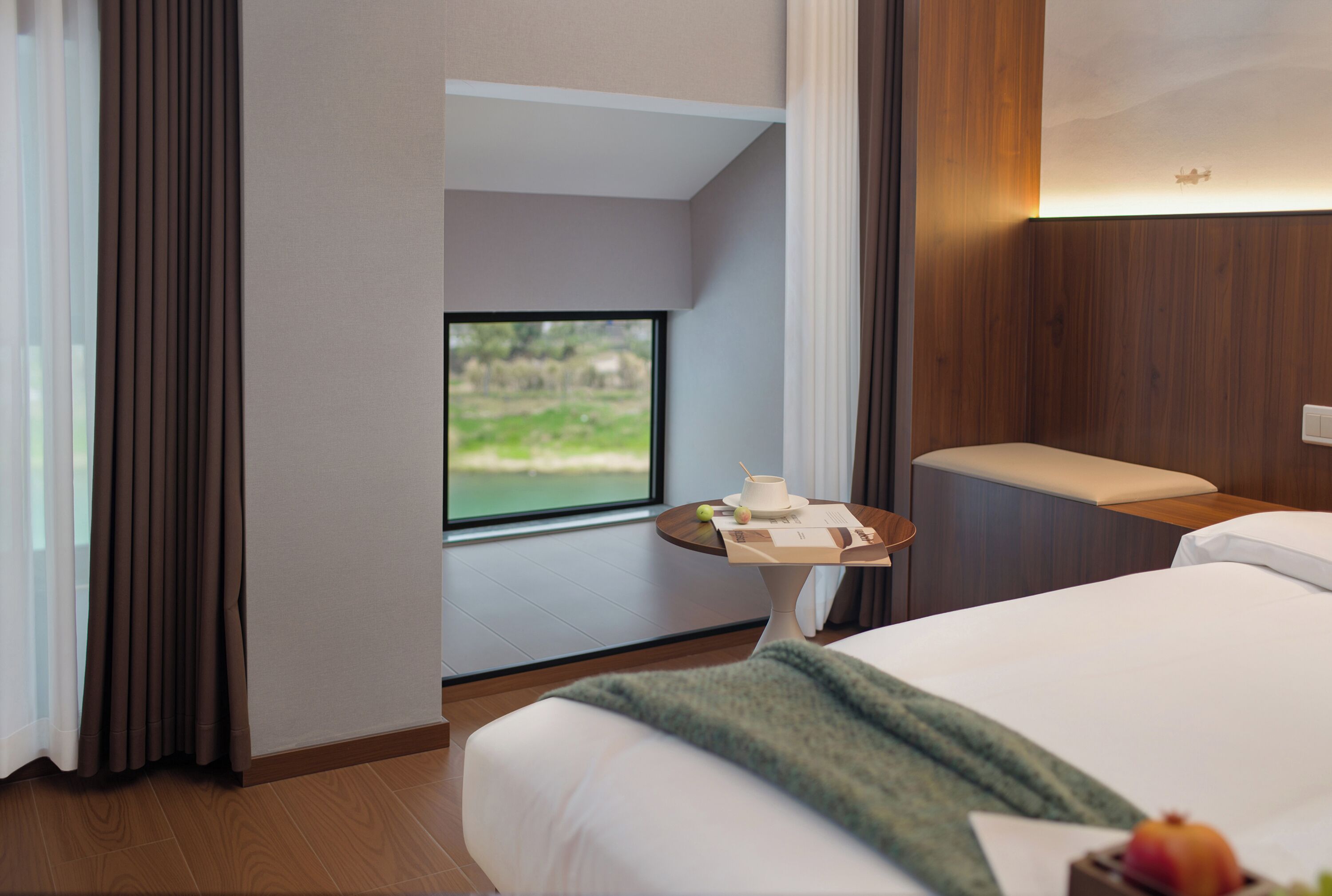 Ein Hotelzimmer mit einem Bett, einem kleinen Tisch mit Tasse und Speisekarte, einem Fenster mit Blick auf Grünflächen und einer Holzwandverkleidung.