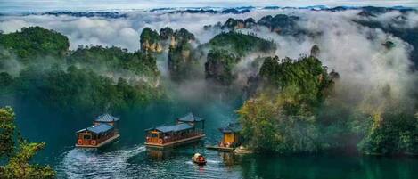 A serene lakeside scene with traditional wooden boats and a small pavilion, surrounded by lush greenery and mist-covered cliffs.