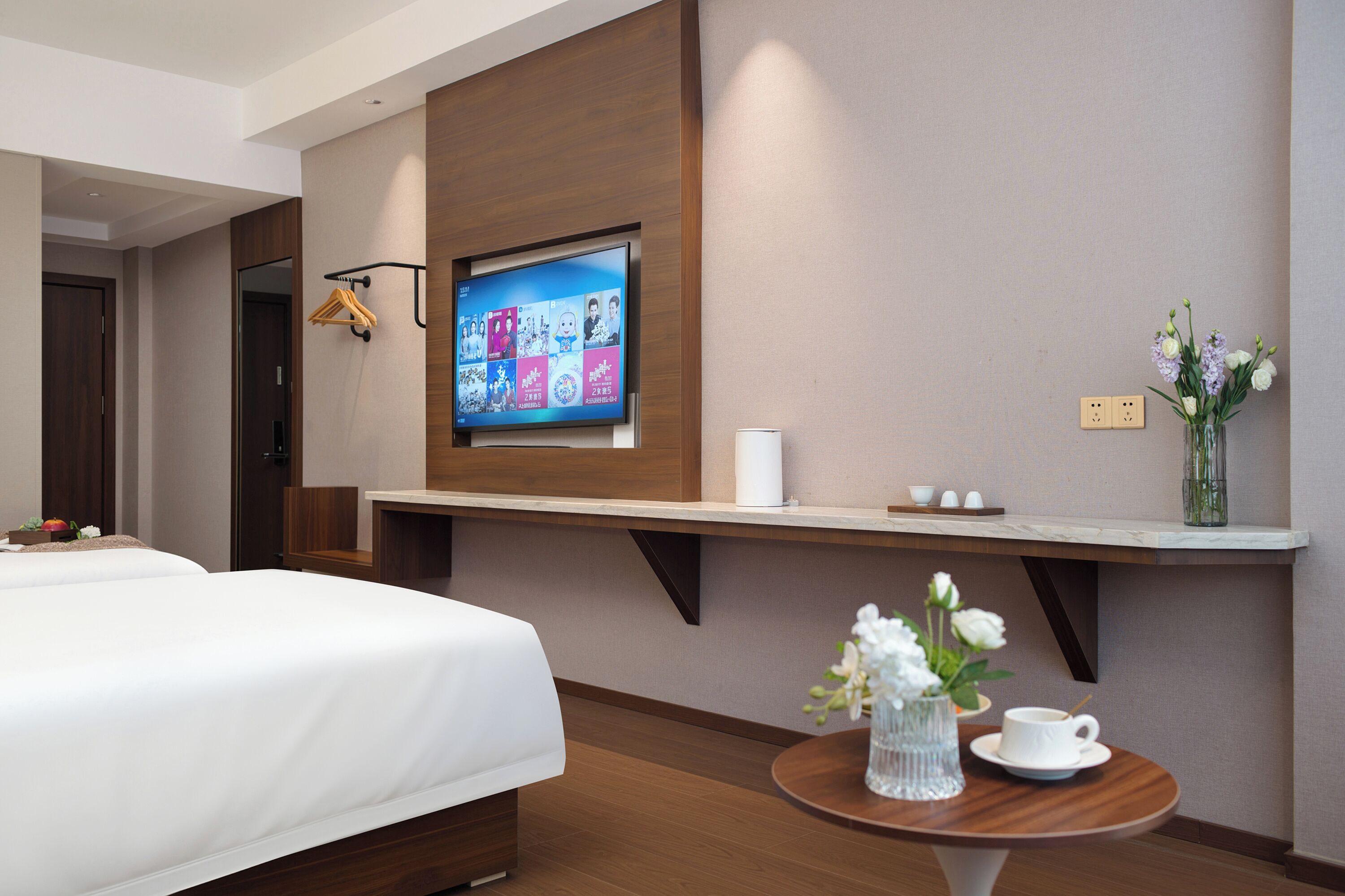 A hotel room with a bed, a TV mounted on the wall, a small table with a vase of flowers, and a shelf with a cup and a tissue.
