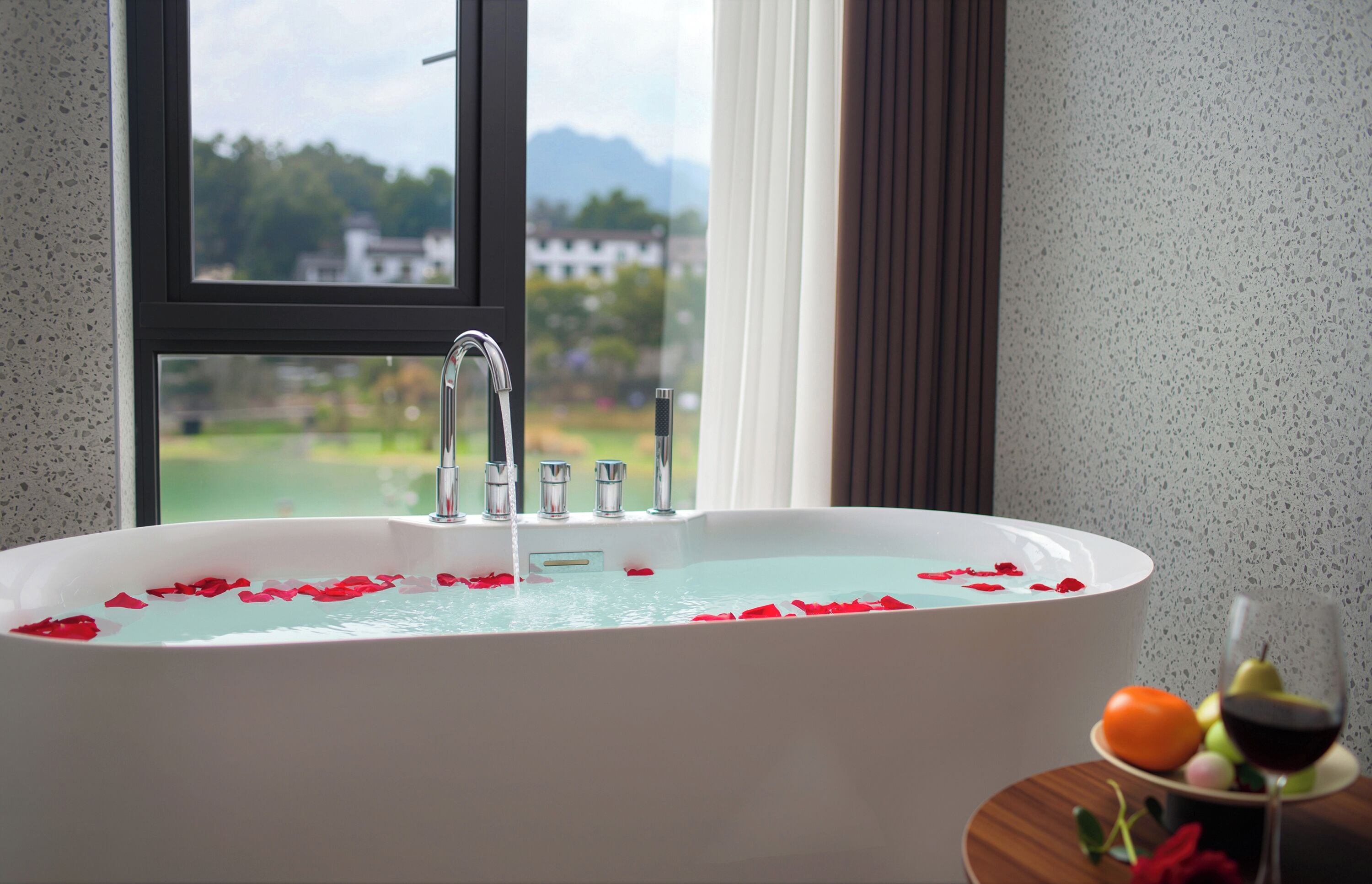 Una bañera con pétalos de rosa, una ventana con vistas a zonas verdes y edificios, y una mesa con una copa de vino y fruta.