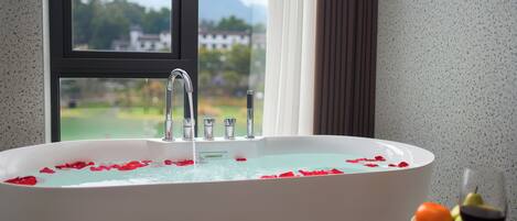 Una bañera con pétalos de rosa, una ventana con vistas a zonas verdes y edificios, y una mesa con una copa de vino y fruta.
