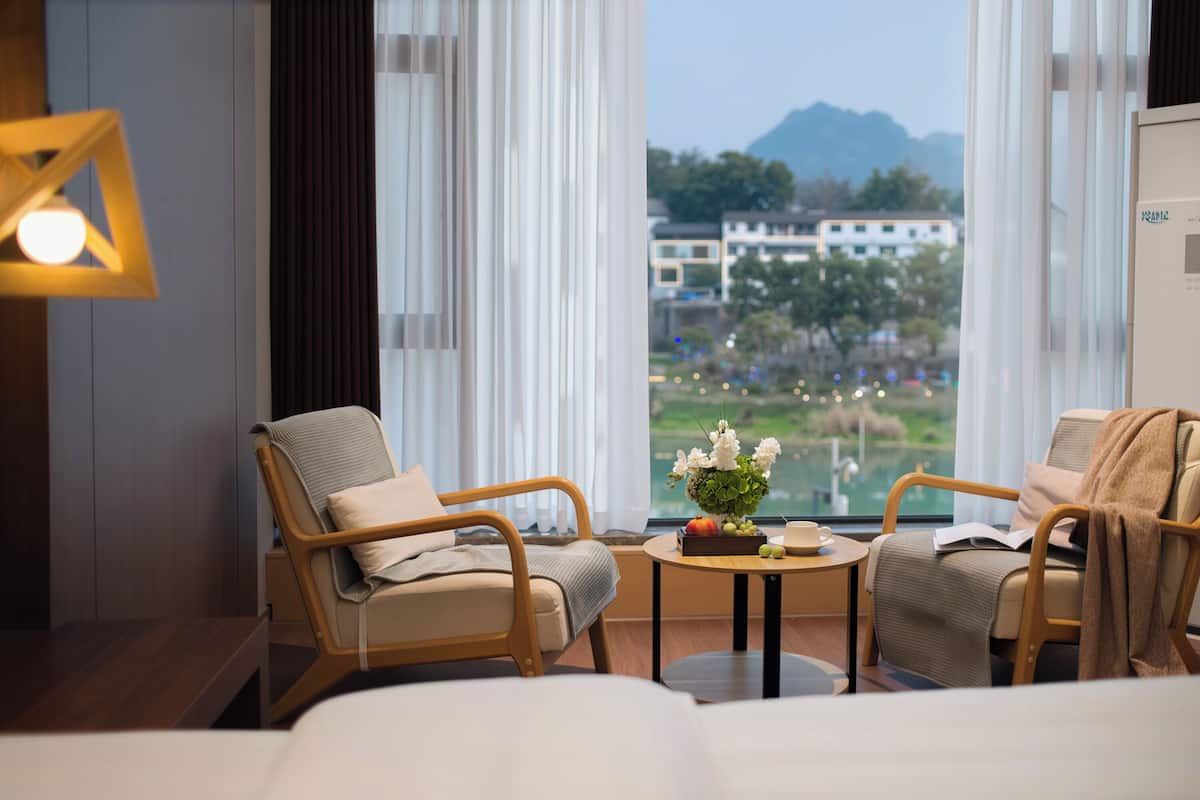 Ein Hotelzimmer mit zwei Stühlen, einem kleinen Tisch mit Blumenvase und einem großen Fenster mit Blick auf Gebäude und Grünflächen.