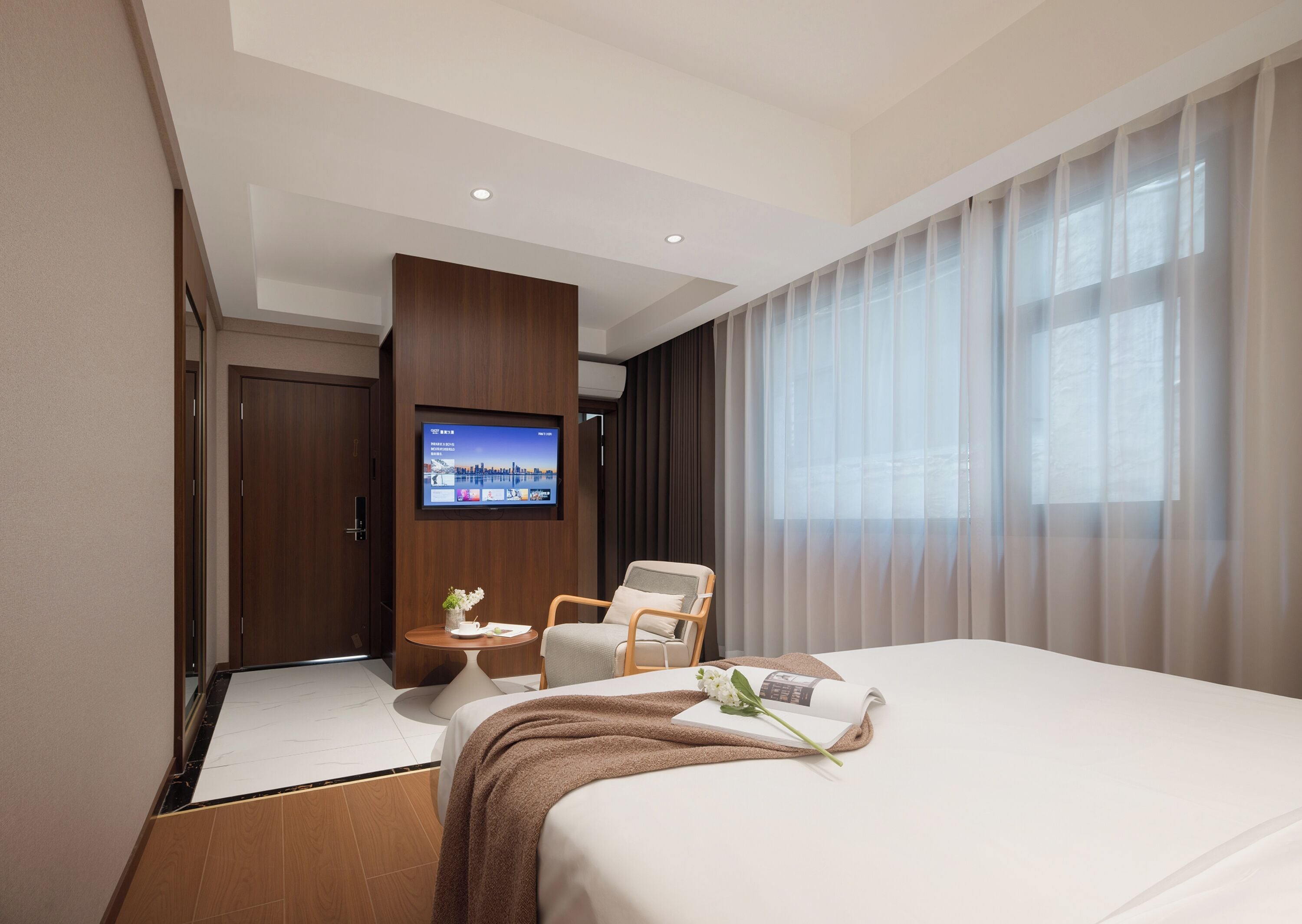 A hotel room with a bed, a TV mounted on the wall, a chair, a small table, and a window with curtains.
