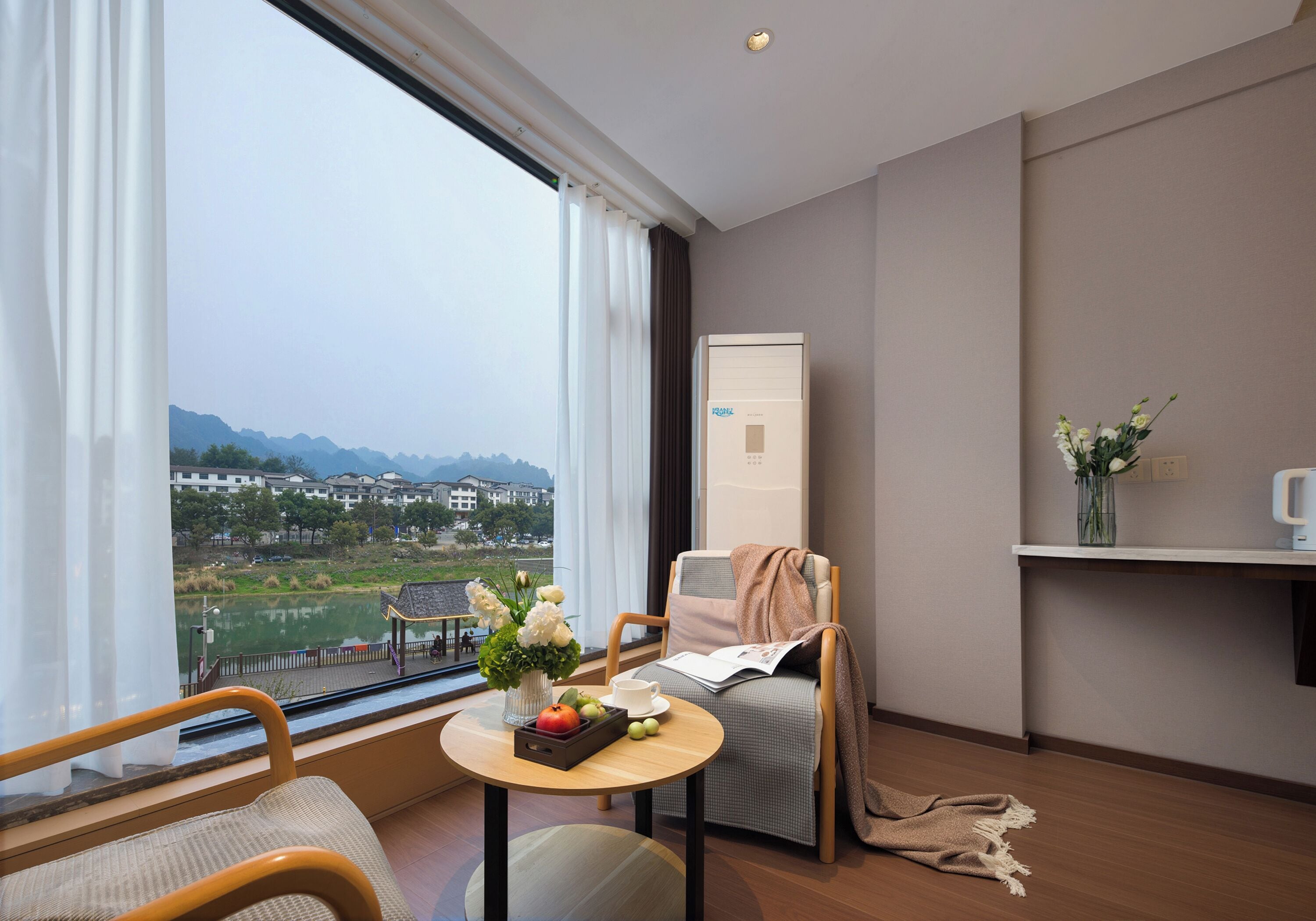 Una habitació d'hotel amb una finestra gran, una cadira, una taula petita amb un ram de flors, una safata amb fruita i vista a un paisatge llunyà.