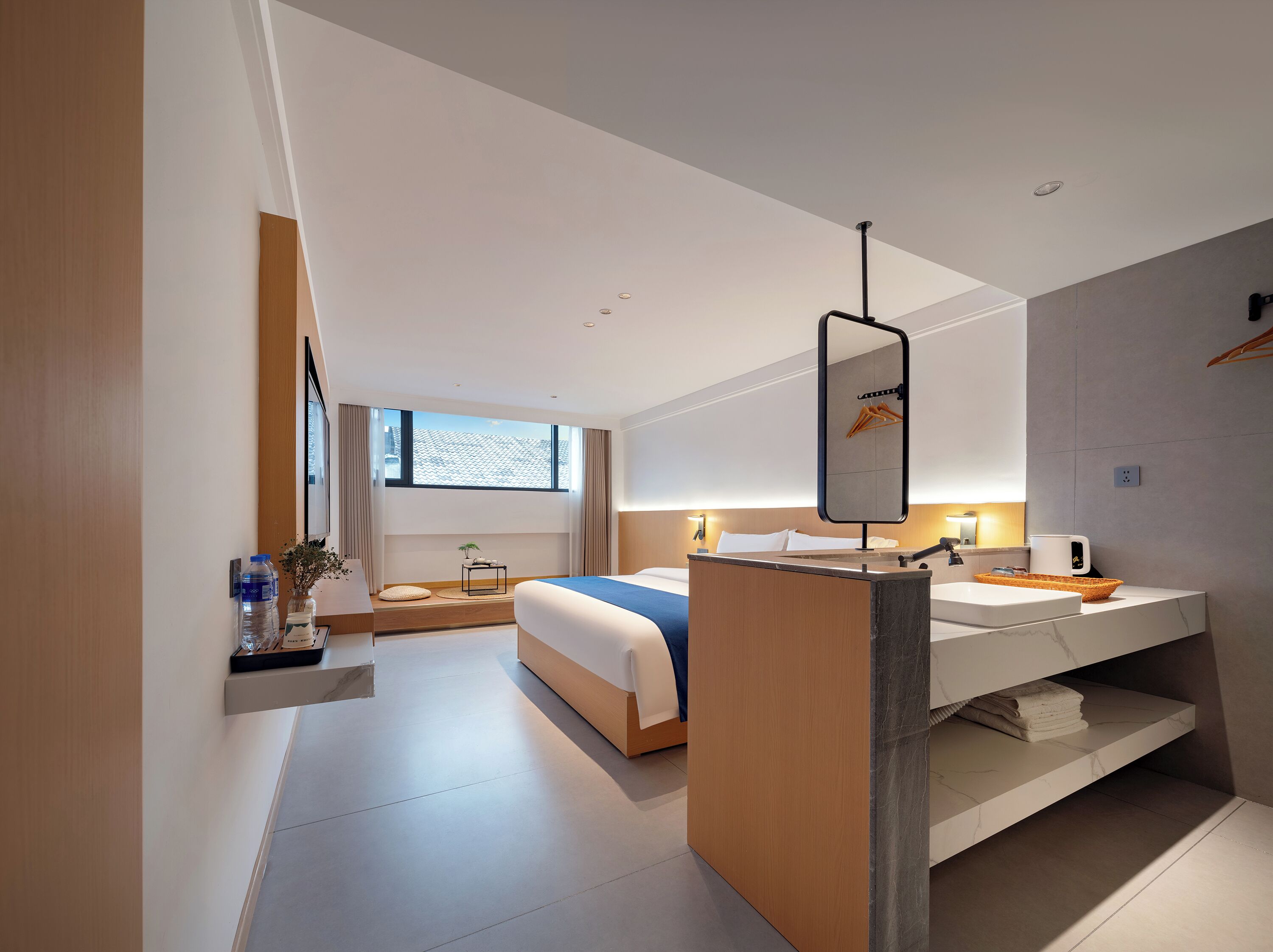 A modern hotel room with a bed, a bedside table, a mirror, a built-in vanity, and a large window.