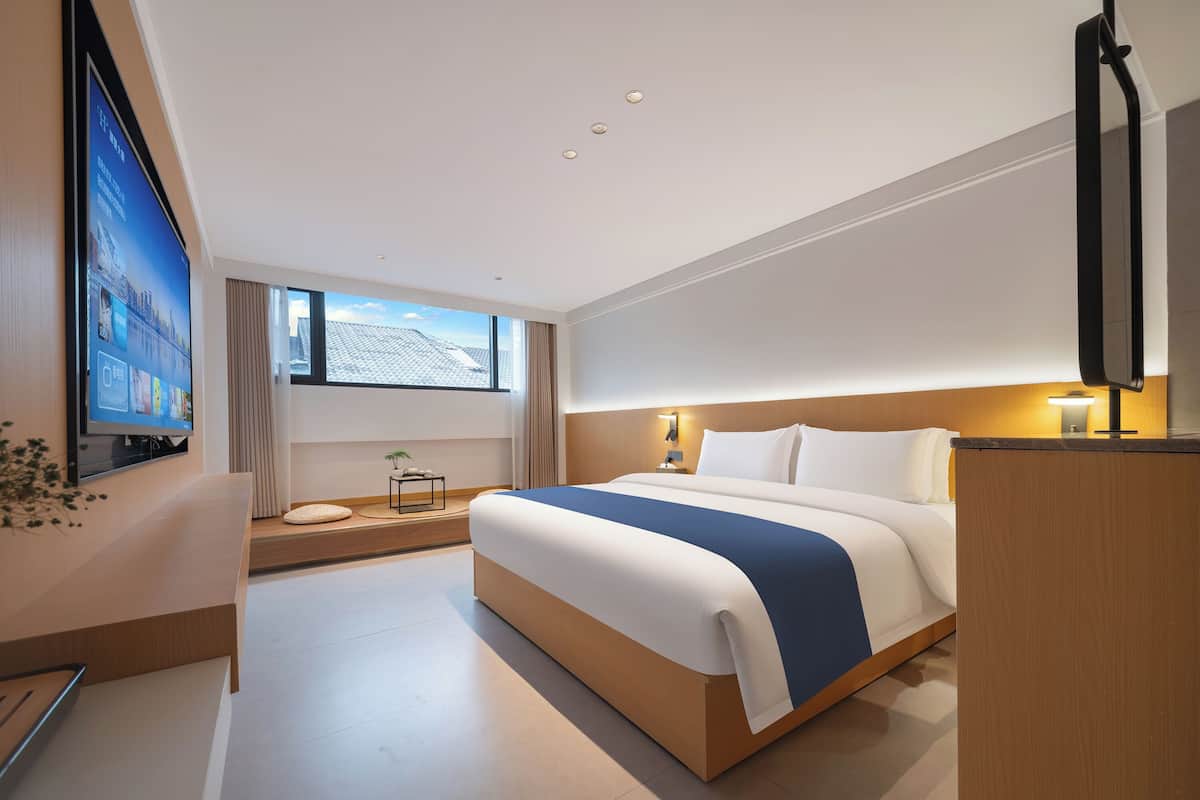 Ein modernes Hotelzimmer mit einem großen Bett, einem an der Wand befestigten Fernseher, einem kleinen Nachttisch mit Lampe und einem Fenster mit Vorhängen.