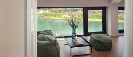 A modern room with a large window, a green bean bag chair, a small table with a vase of flowers, and a view of a lake.
