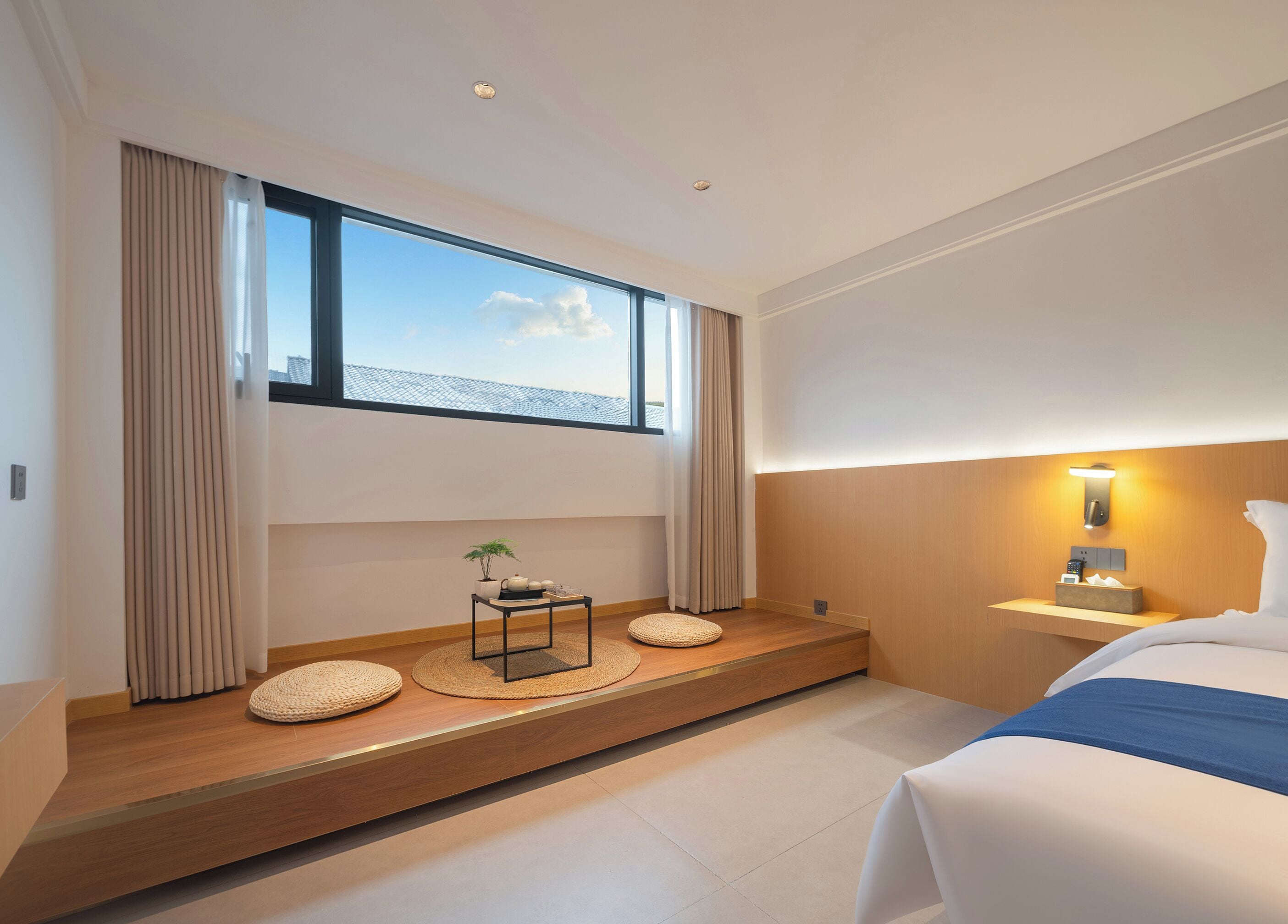 A modern hotel room with a large window, a bedside table with a lamp, and a bed with a blue accent.