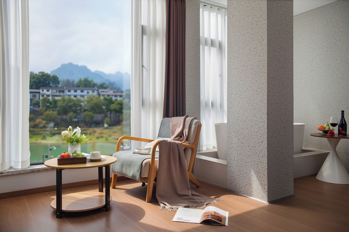 Ein Zimmer mit Bergblick, ein Sessel mit Decke, ein Tisch mit Vase und Buch sowie ein kleiner Tisch mit Obst und einer Flasche.