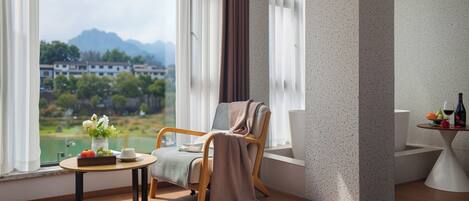 A room with a view of mountains, a chair with a blanket, a table with a vase and a book, and a small table with fruit and a bottle.