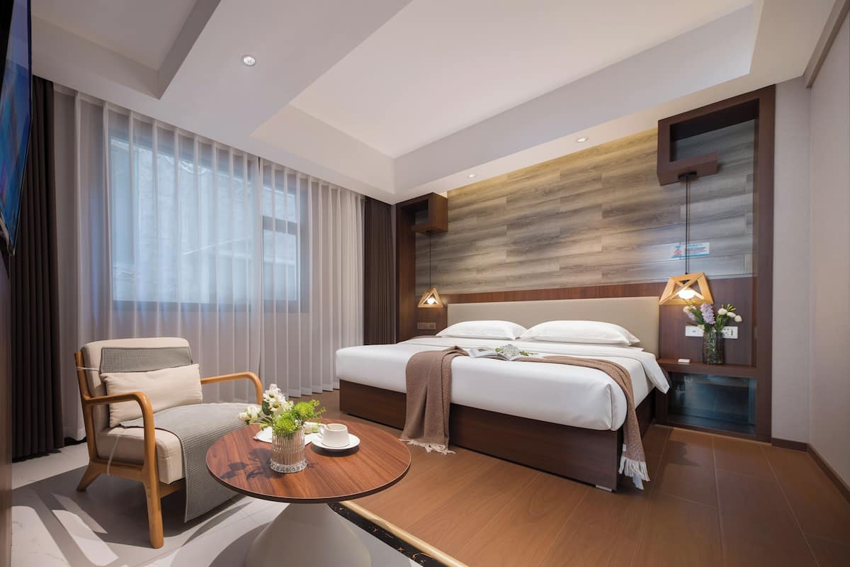 Ein modernes Hotelzimmer mit einem Bett, einem Sessel, einem kleinen Tisch mit einer Vase und einem Nachttisch mit einer Lampe.