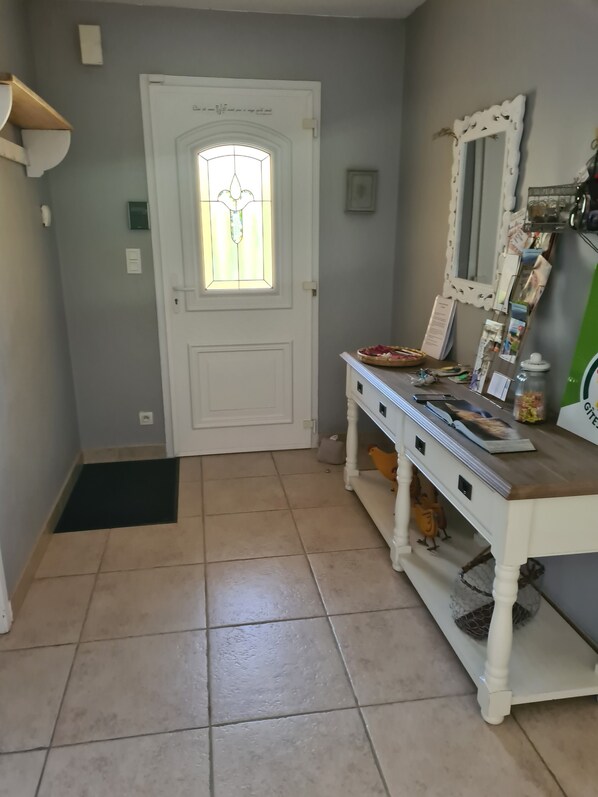 Un pasillo con una puerta blanca, un ventanal pequeño con diseño de vitral, un banco blanco con varios objetos y un piso de baldosas.