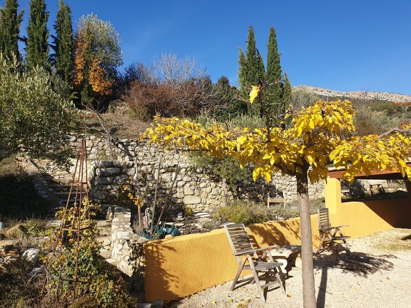A rustic outdoor seating area with stone walls and a yellow fence, surrounded by trees and a clear blue sky.