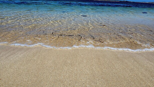 A beach with clear shallow waters, a sandy shore, and a distant rocky outcrop.